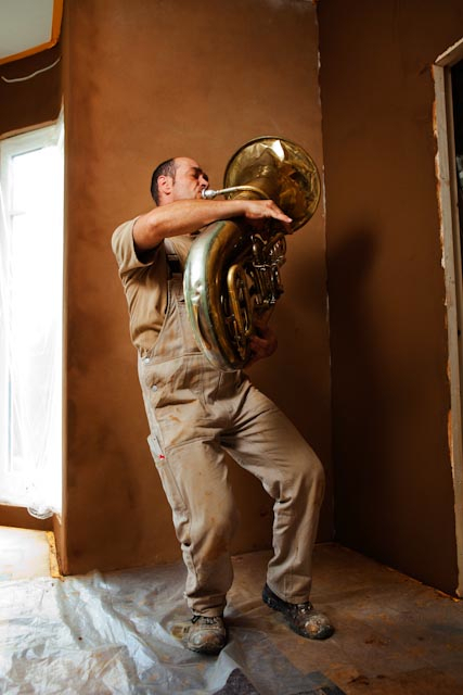 Handwerker spielt Tuba vor Lehmwand